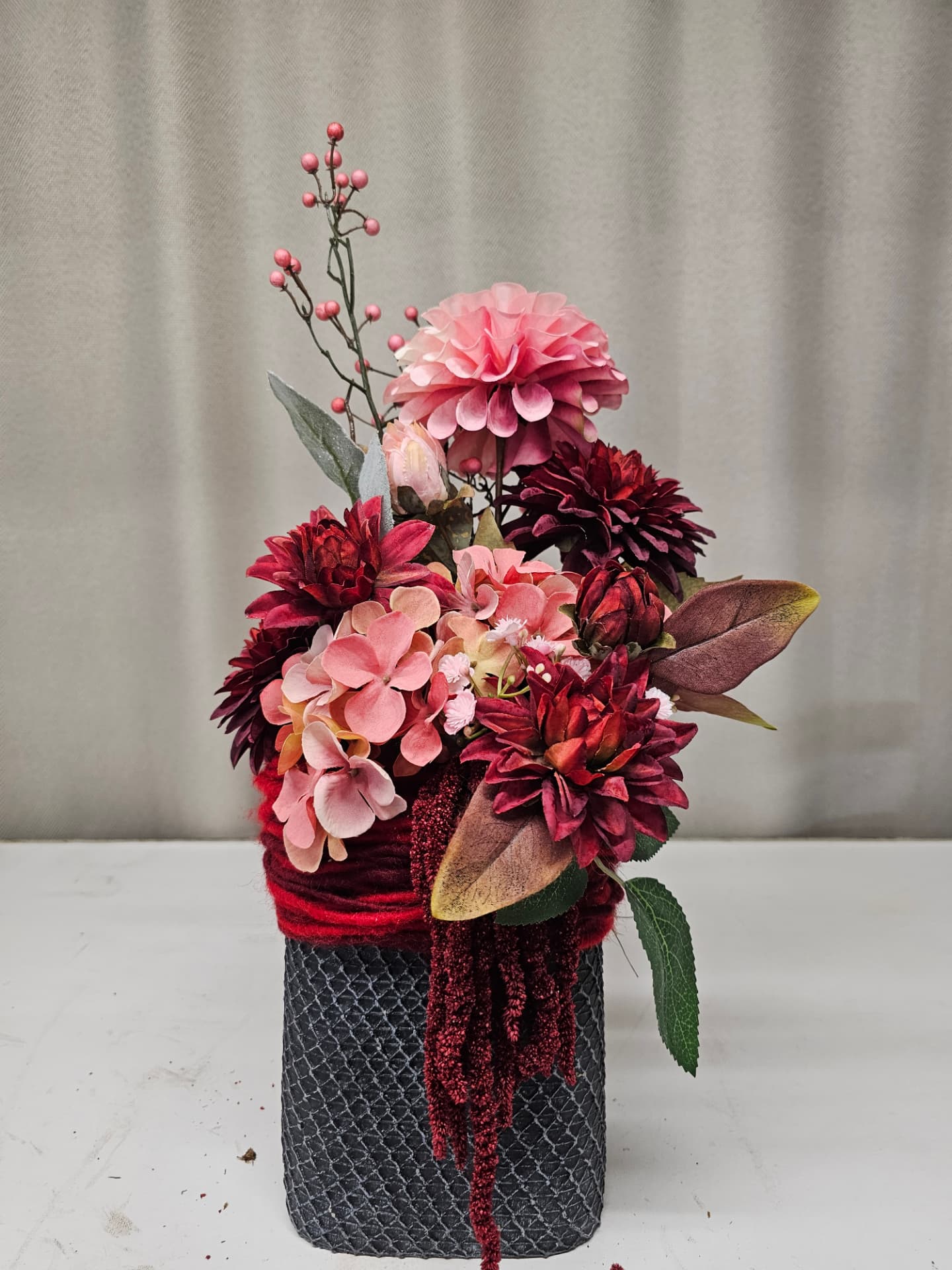 Hoog grafstukje in een donkergrijze pot met rode wollen draad, gevuld met zijdebloemen